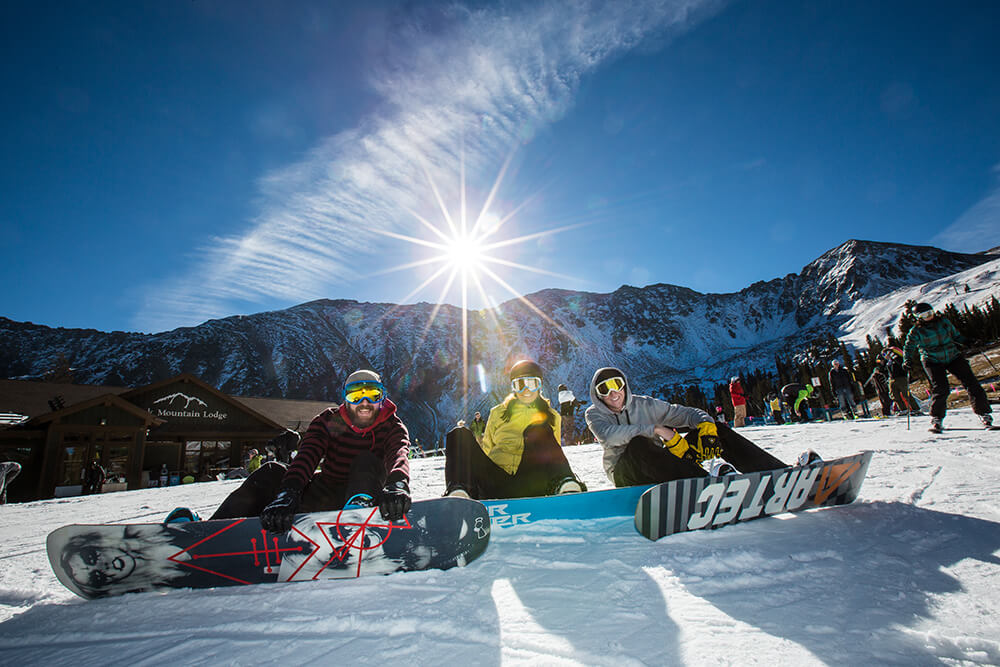 Smiling snowboarders laying on the ground with mountains in the background Arapahoe Basin