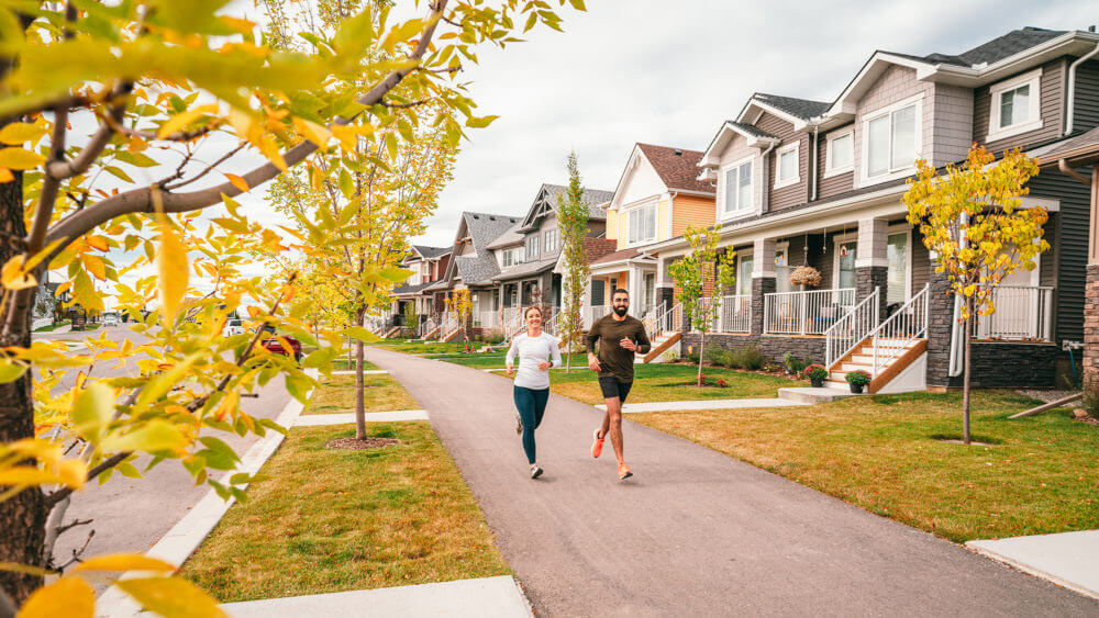 A couple running down a wide sidewalk in Vista Crossing community, passing charming houses, green lawns and trees.