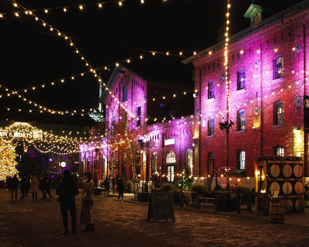 Nighttime image of the distillery district's 2020 Winter Village celebration with Christmas lights hanging over the space and people walking by