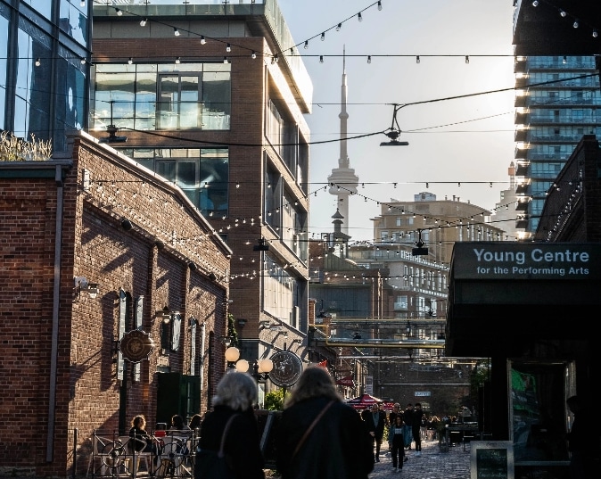 A summer scene in the Distillery district: people strolling down a brick alleyway with buildings and the Cn Tower in the background, and the Young Centre for the Performing Arts stands in the forefront.