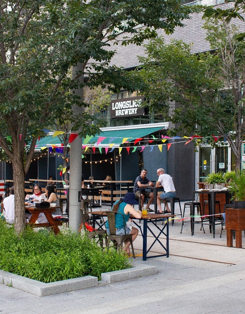 a street view on Longslice Breweries patio sits underneath green trees, and rainbow colored flags as people enjoy each other's company.