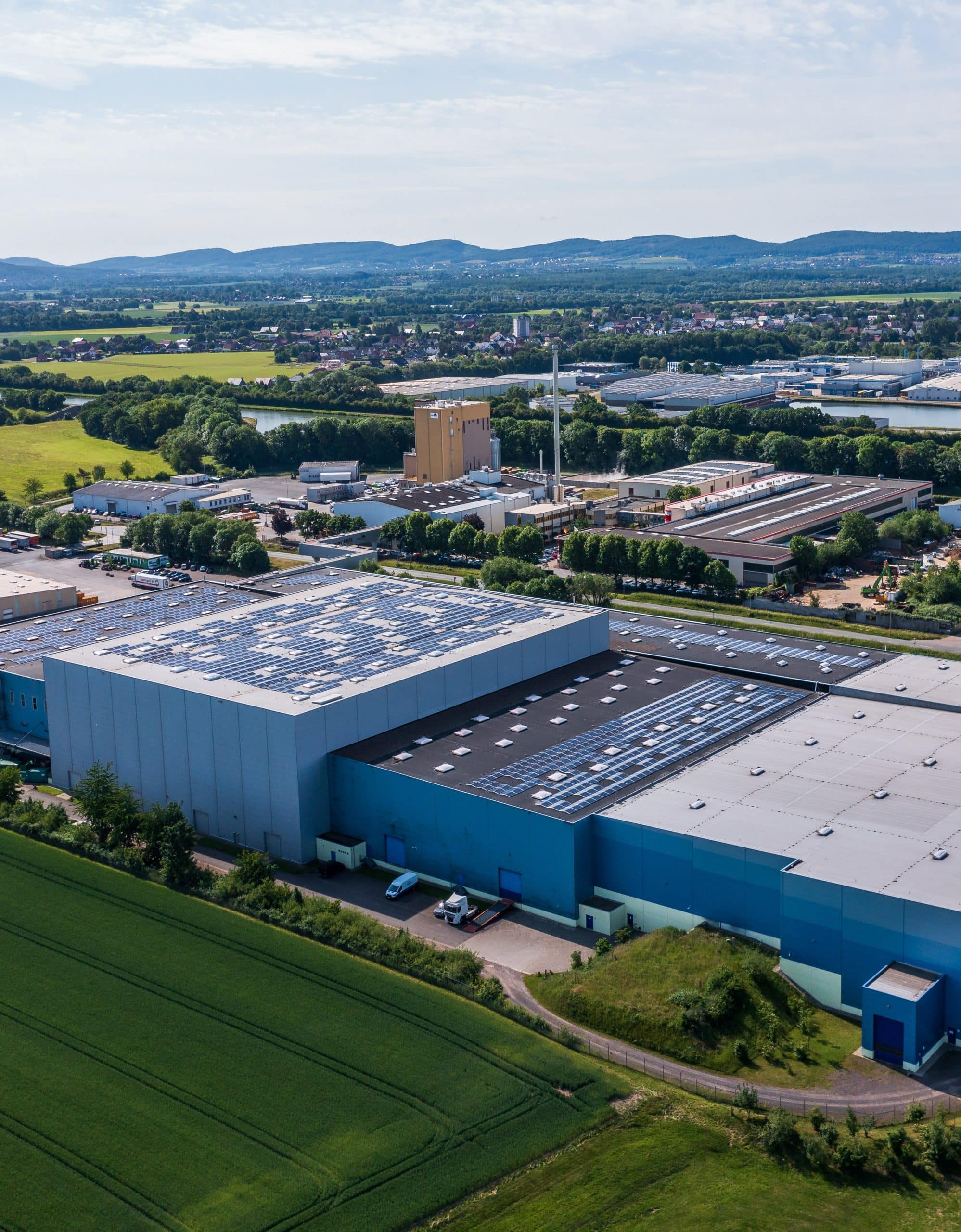 Aerial view of a large industrial complex in Germany, with a blue building featuring rooftop solar panels, surrounded by green fields, smaller structures, a tall beige building with a chimney, and distant residential areas under a partly cloudy sky