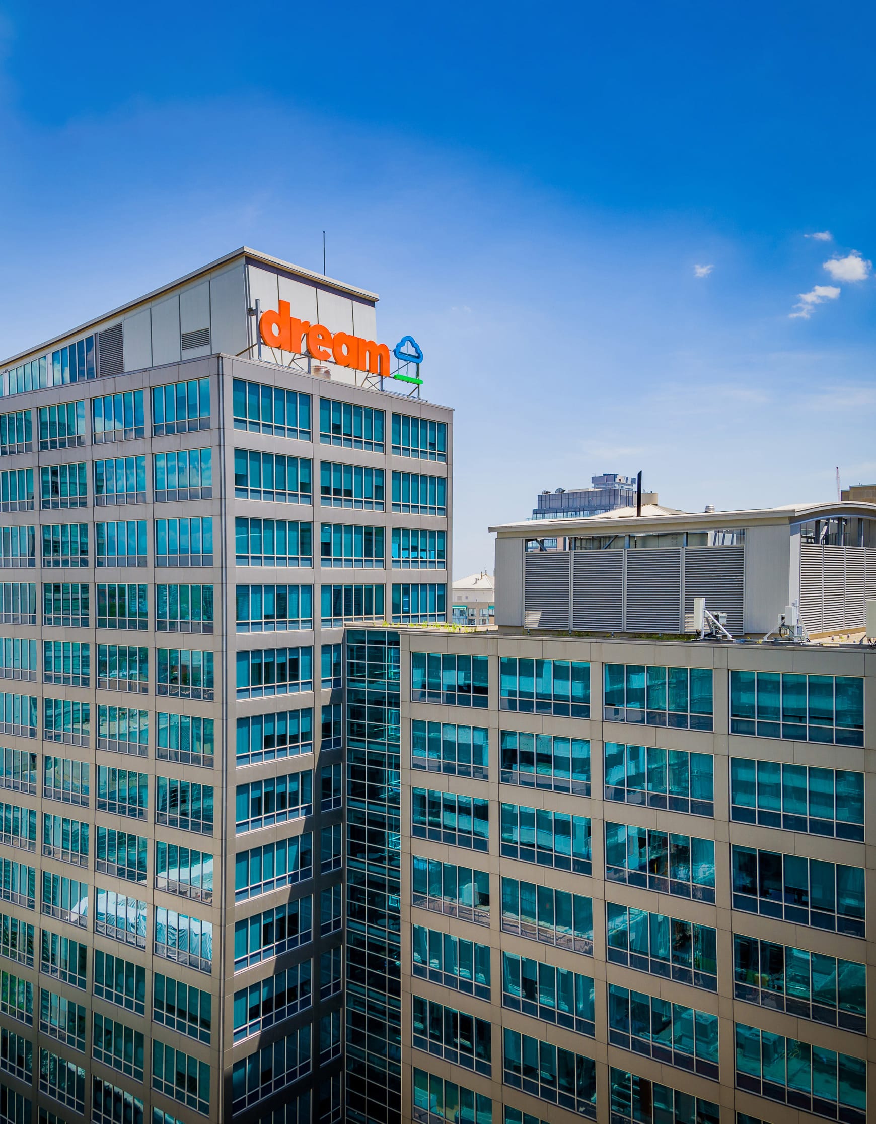 Modern multi-story office building with large glass windows and a rooftop sign that reads "dream" in orange letters next to a blue cloud icon, set against a clear blue sky on a sunny day