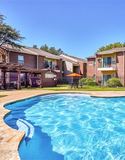 Outdoor swimming pool with rippling water and a stone deck, surrounded by two-story brick apartment buildings with balconies; a pergola stands to the left and a picnic table with an orange umbrella sits near the center, under a clear blue sky