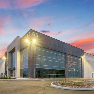 Modern industrial or commercial building with large glass windows and the number '65' displayed on the upper corner, surrounded by small trees and landscaping, with a paved driveway in front and a colorful sky suggesting sunrise or sunset